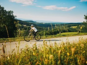 Eine Person f&auml;hrt mit einem Fahrrad auf einer l&auml;ndlichen Stra&szlig;e, im Hintergrund weiter Aussicht.