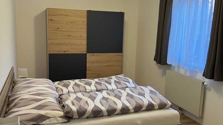 Ein Schlafzimmer mit einem Doppelbett, einem Kleiderschrank und einem Nachttisch. Die W&auml;nde sind wei&szlig;, der Boden ist aus Holz und ein Fenster mit Vorh&auml;ngen ist zu sehen.