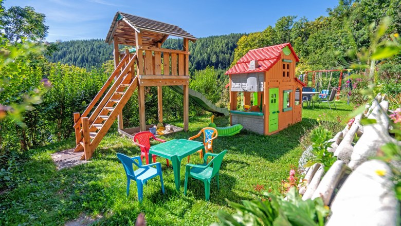 Ein bunter Spielplatz mit Klettergerüst, Rutsche, Spielhaus und Tisch mit Stühlen auf einer Wiese, umgeben von Bäumen und Bergen.