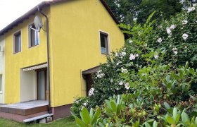 Gelbes Haus mit Terrasse und blühenden Sträuchern im Vordergrund.