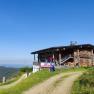 Eine Berghütte aus Holz mit einem roten Fahrzeug davor, umgeben von grünen Wiesen und einem klaren blauen Himmel.