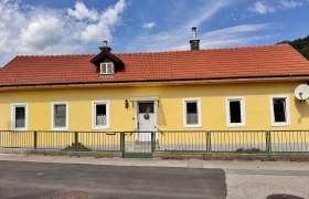 Gelbes Haus mit rotem Dach und mehreren Fenstern, umgeben von einem gr&uuml;nen Zaun.