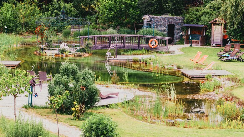 Ein idyllischer Garten mit Teich, Holzbr&uuml;cke und Liegest&uuml;hlen.