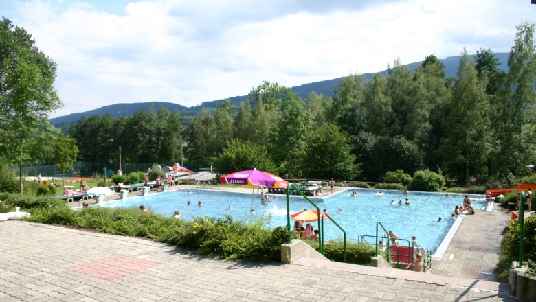 Ein Freibad mit Menschen im Wasser, umgeben von B&auml;umen und Bergen im Hintergrund.