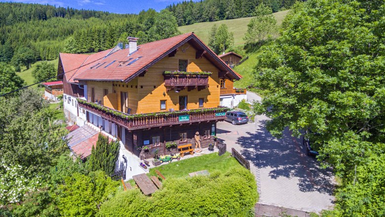 Ein traditionelles Holzhaus in einer gr&uuml;nen, h&uuml;geligen Landschaft mit B&auml;umen und einem blauen Himmel.