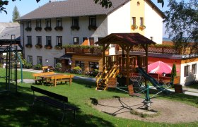 Ein Gasthof mit Spielplatz im Vordergrund, umgeben von gr&uuml;ner Wiese und B&auml;umen.