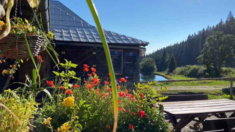 Blumen vor einer Bergh&uuml;tte mit Teich und Wald im Hintergrund.