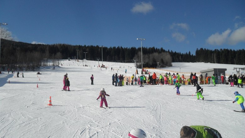 Kinder und Erwachsene beim Skifahren auf einer Piste mit blauem Himmel.