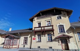 Historisches Bahnhofsgebäude mit gelber Fassade und rotem Holzdekor unter blauem Himmel.