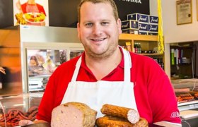 Ein Metzger in rotem Hemd und wei&szlig;er Sch&uuml;rze h&auml;lt verschiedene Wurstwaren in einer Fleischerei.