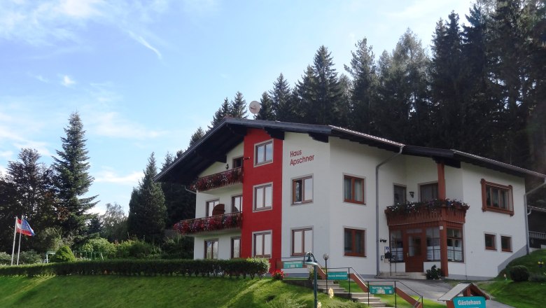 Ein zweist&ouml;ckiges G&auml;stehaus mit roten Balkonen und Blumen, umgeben von B&auml;umen und einem blauen Himmel.