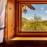 Ausblick aus kleinem Holzfenster mit wei&szlig;em Vorhang auf B&auml;ume unter blauem Himmel.