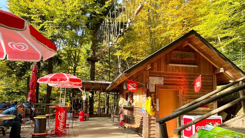 Holzhütte mit Sonnenschirmen und Kletterpark im Hintergrund.