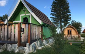 Zwei kleine Holzh&uuml;tten im Glamping-Stil mit gr&uuml;ner und brauner Fassade, umgeben von B&auml;umen und Gras.