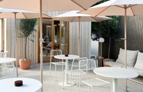 Moderne Terrasse mit wei&szlig;en Tischen und St&uuml;hlen, Sonnenschirmen und Holzh&uuml;tte im Hintergrund.