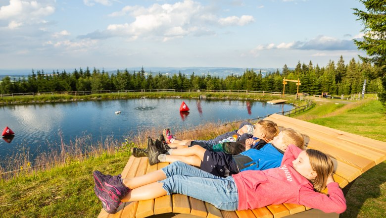 Kinder entspannen auf einer Holzbank am Speicherteich in St. Corona am Wechsel.