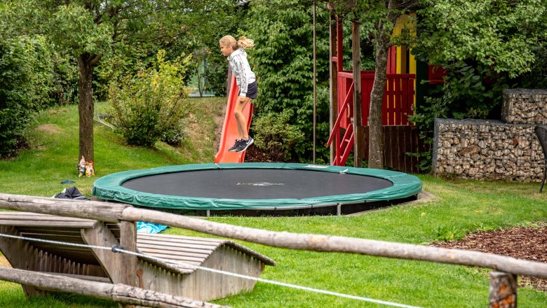Ein Kind springt auf einem Trampolin in einem Garten mit Rutsche und Spielhaus.