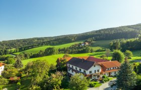 Ländliche Landschaft mit einem großen Haus, umgeben von grünen Feldern und Bäumen unter blauem Himmel.