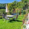 Ein Spielplatz mit Tisch, Stühlen, Sonnenschirm und Schaukeln auf einer Wiese, umgeben von Bäumen und Blumen.