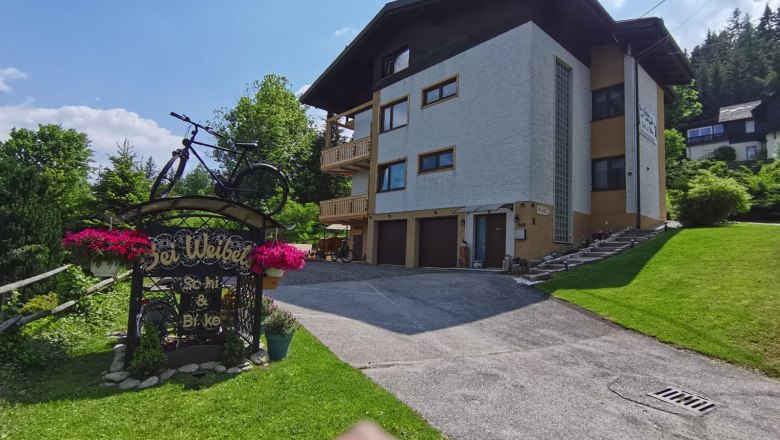 Ein gro&szlig;es Haus mit mehreren Etagen, umgeben von gr&uuml;ner Landschaft. Im Vordergrund ein Schild mit einem Fahrrad und Blumen.