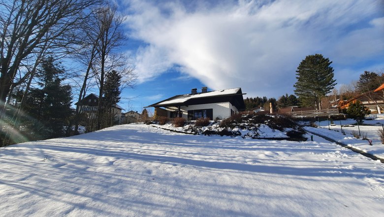 Winterlandschaft mit einem Haus im Schnee, umgeben von Bäumen und blauem Himmel.