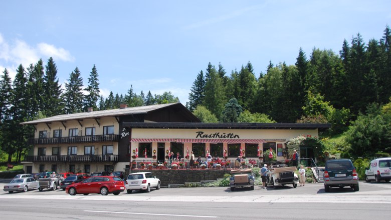Ein Rasthaus mit Terrasse und geparkten Autos davor, umgeben von Bäumen.