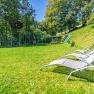 Spielplatz mit Rutsche, Trampolin und Liegestühlen auf einer Wiese, umgeben von Bäumen.