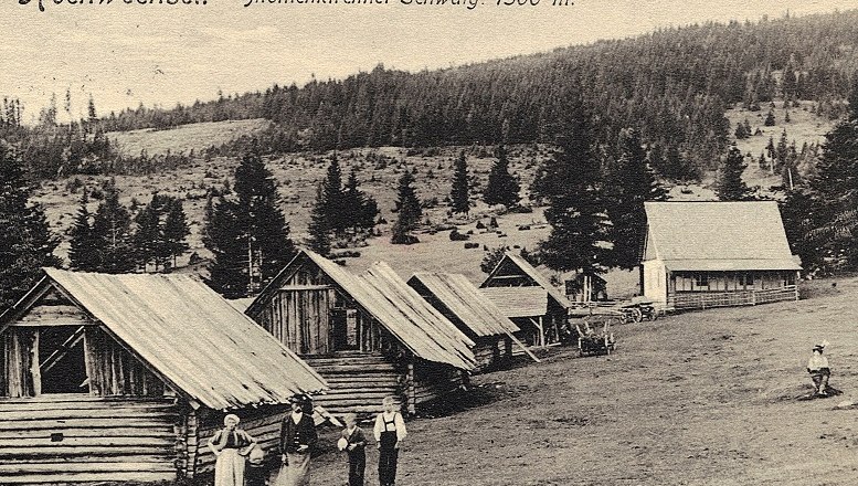 Historische Aufnahme von Holzhütten auf der Mönichkirchner Schwaig, umgeben von Wald und Wiesen, mit Menschen im Vordergrund.