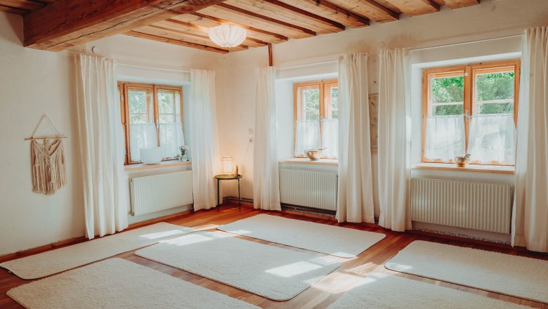 Ein heller Yogaraum mit Holzboden, weißen Matten und großen Fenstern mit Vorhängen.