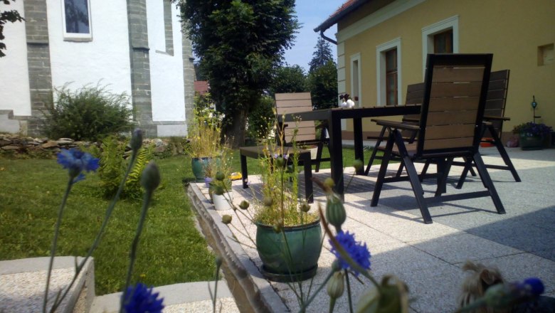 Terrasse mit Tisch, Stühlen und Pflanzen in einem Garten neben einem gelben Haus.