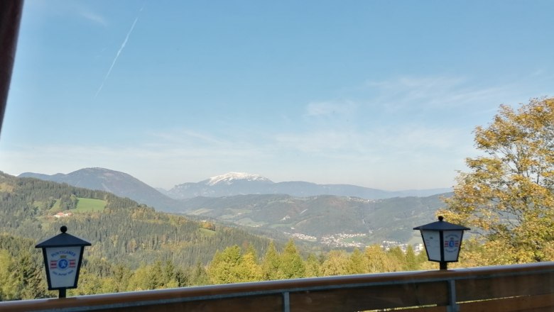 Blick von einem Restaurantbalkon auf eine bergige Landschaft mit Wald und einem schneebedeckten Gipfel im Hintergrund.
