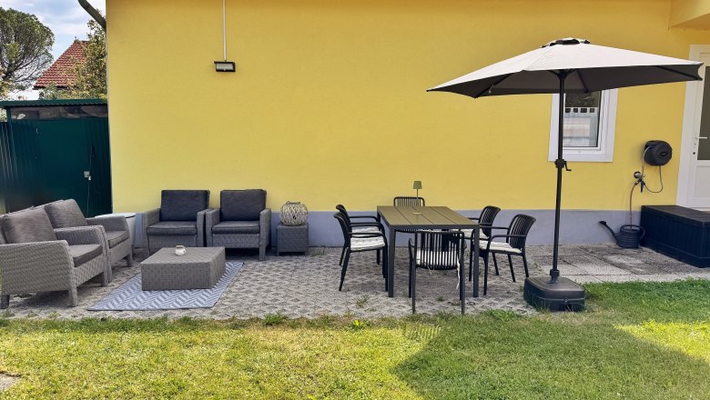 Terrasse mit gelber Hauswand, grauen Sitzm&ouml;beln, Esstisch, St&uuml;hlen und Sonnenschirm.