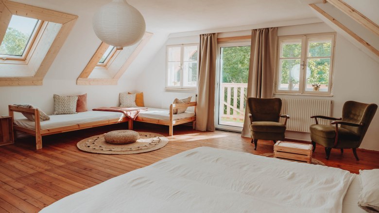 Helles Mehrbettzimmer mit Holzboden, zwei Betten, zwei Sesseln und großen Fenstern.