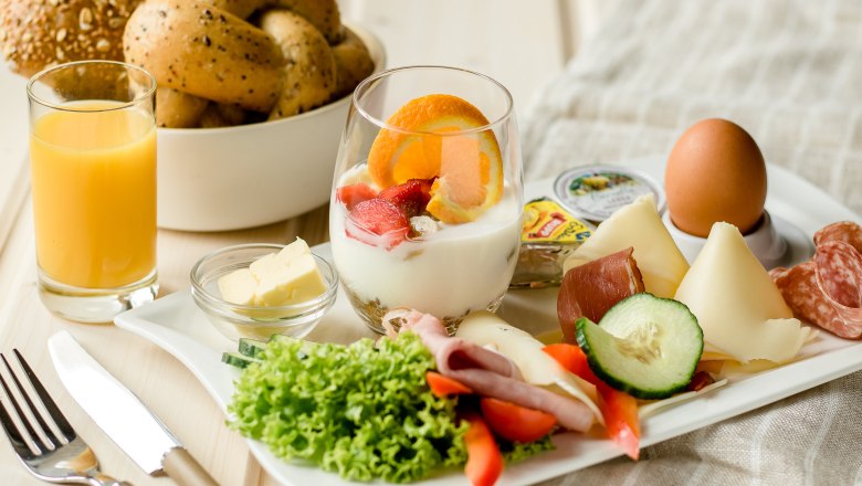 Ein Fr&uuml;hst&uuml;ckstablett mit Joghurt, Obst, K&auml;se, Wurst, Ei, Salat, Butter und Orangensaft.