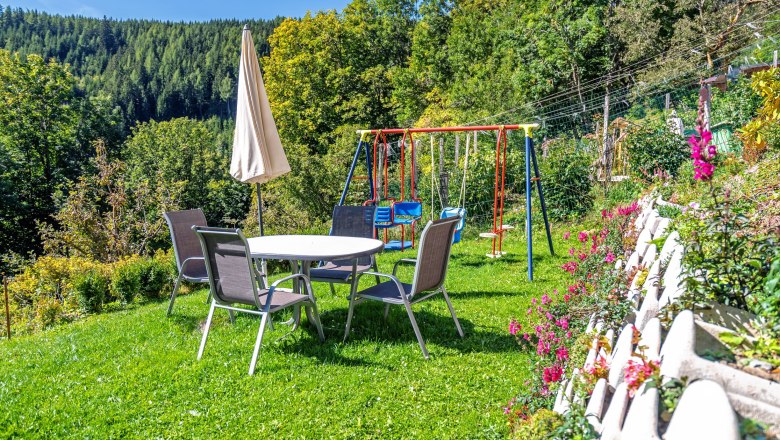 Ein Spielplatz mit Tisch, Stühlen, Sonnenschirm und Schaukeln auf einer Wiese, umgeben von Bäumen und Blumen.
