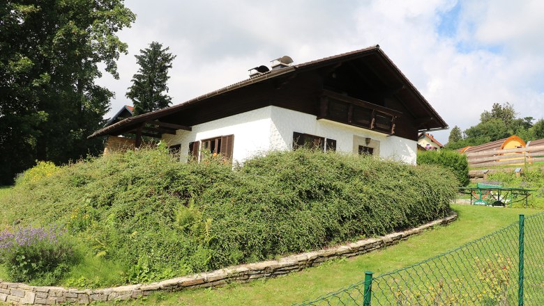 Ein wei&szlig;es Haus mit Holzdach und Garten, umgeben von B&uuml;schen und B&auml;umen, unter einem bew&ouml;lkten Himmel.