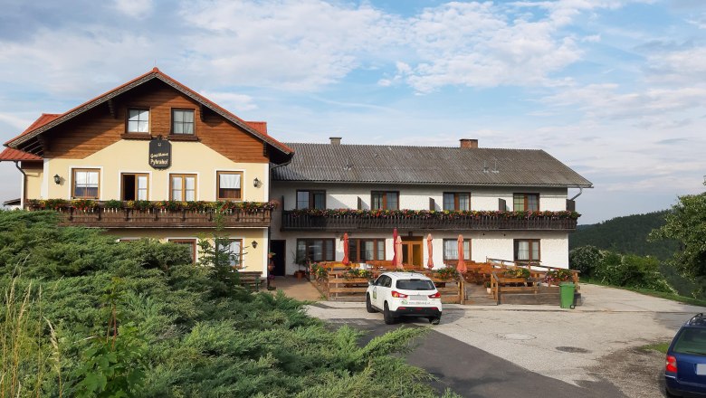 Ein traditionelles Gasthaus mit Balkon und Blumenk&auml;sten, davor ein Parkplatz mit Autos, umgeben von gr&uuml;ner Landschaft und blauem Himmel.