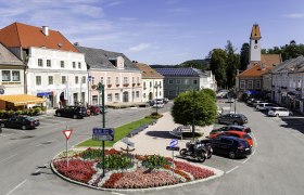 Hauptplatz in Aspang Markt mit bunten Blumenbeeten, geparkten Autos und historischen Geb&auml;uden.