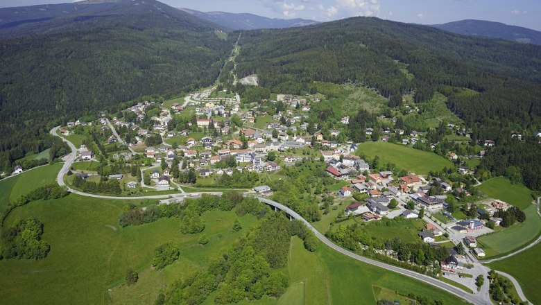 Luftaufnahme eines Dorfes in einer gr&uuml;nen, h&uuml;geligen Landschaft mit W&auml;ldern und Wiesen.