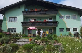 Ein grünes Haus mit Balkonen und Menschen davor, umgeben von einem blühenden Garten.