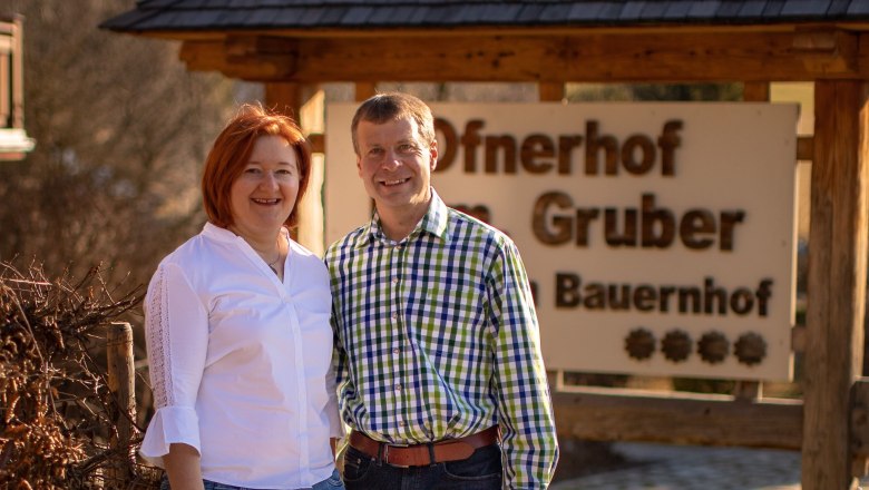 Ein Mann und eine Frau stehen l&auml;chelnd vor einem Schild mit der Aufschrift 'Ofnerhof Gruber Bauernhof'.