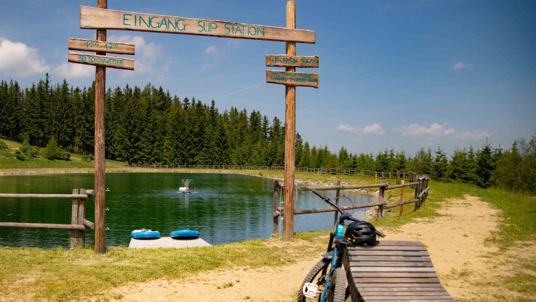 Speicherteich mit SUP-Station in St. Corona am Wechsel, umgeben von Wald und einem Fahrrad im Vordergrund.