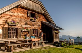 Holzh&uuml;tte in den Bergen mit Menschen am Tisch, blauer Himmel.
