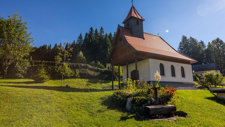 Kleine Kapelle mit rotem Dach auf einer gr&uuml;nen Wiese, umgeben von B&auml;umen und Blumen.