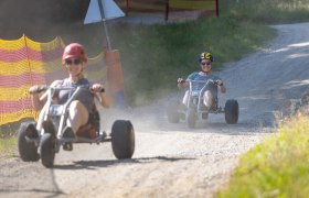 Zwei Personen fahren auf Schotterweg mit Mountaincarts, tragen Helme und Sonnenbrillen.