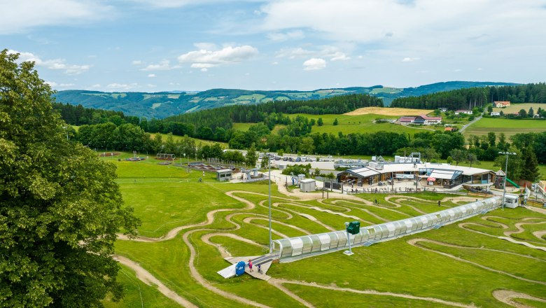 Luftaufnahme der Wexl Arena in St. Corona am Wechsel mit gr&uuml;nen H&uuml;geln und einem Geb&auml;ude im Hintergrund.