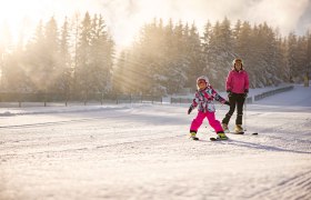 Kind und Erwachsener beim Skifahren im Schnee, umgeben von Bäumen und Sonnenstrahlen.