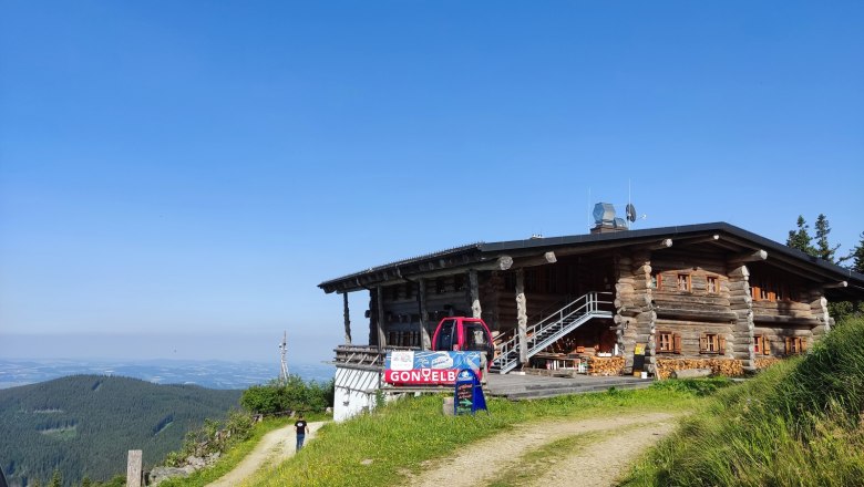Eine Berghütte aus Holz mit einem roten Fahrzeug davor, umgeben von grünen Wiesen und einem klaren blauen Himmel.