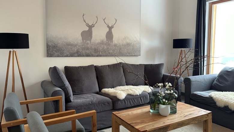 Wohnzimmer mit grauen Sofas, Holztisch und Hirschbild an der Wand.