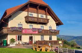 Ein traditionelles Gasthaus mit Holzbalkonen und Blumen, umgeben von gr&uuml;nen H&uuml;geln und blauem Himmel.